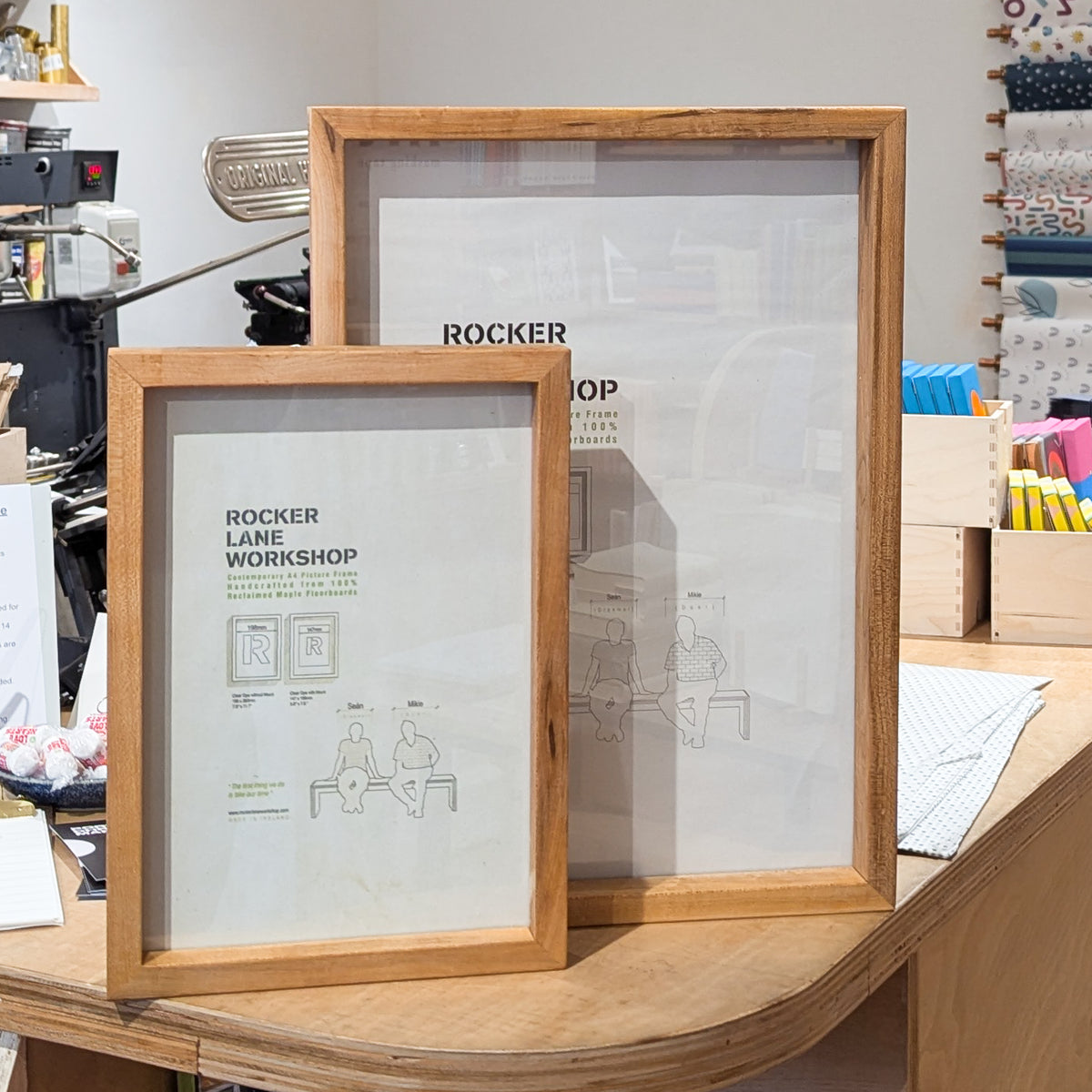 Two wooden frames with glass panes, one larger and one smaller, displaying a diagram and text related to reclaimed maple floorboards, placed on a counter in a store.